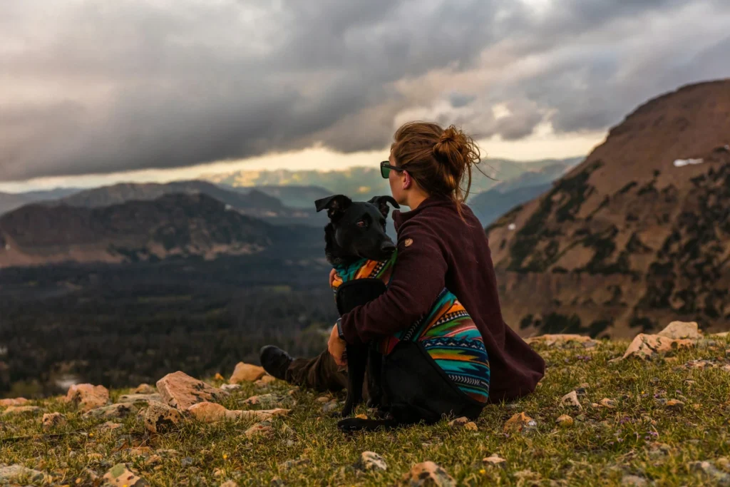 A woman sitting on a grassy mountain peak hugging her black dog.